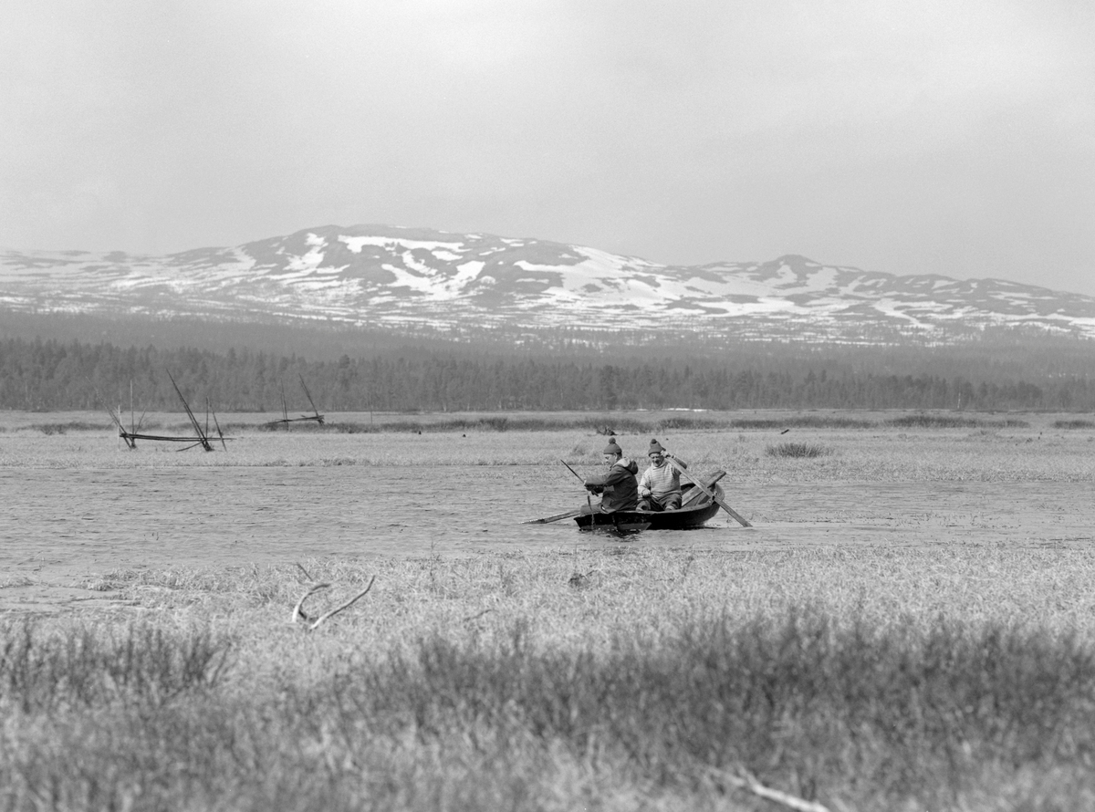 Gjeddefiske i deltaområdet der elva Tufsinga renner ut i innsjøen Femund. Lokaliteten ligger i Tufsingdalen i Os kommune i Nord-Østerdalen, bortimot 700 meter over havet. Fotografiet viser to menn i en klinkbygd trebåt med tverr akterende. Karene skulle trekke gjeddegarn. Vi vet foreløpig ikke hvem mannen i akterenden av båten var. Han som satt ved årene var «gjeddekongen» Joseph Bakken (1898-1989). Gjeddefisket i dette området foregikk i mai måned, like etter at isen på vassdragene hadde smeltet. Da trakk gjedda inn mot de sivbevokste grunnene langs land for å gyte. Der satte fiskerne garn skrått ut fra strandlinja. Det var slike garn de to mennene i båten skulle sette eller trekke da dette fotografiet ble tatt.