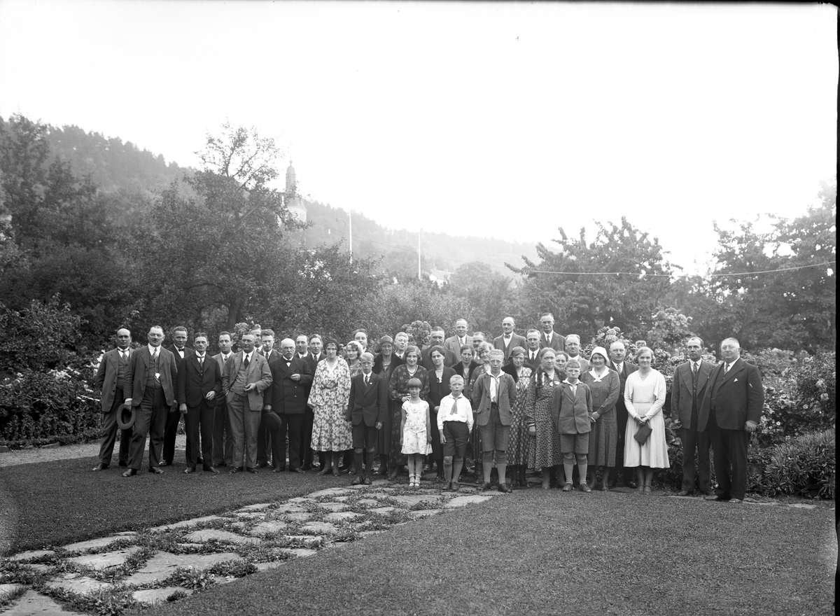 En större grupp med kvinnor, män och barn i Ribbagårdens trädgård. Gränna kyrka anas till vänster i bakgrunden.