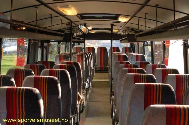 Turkis buss med en sort stripe som går rundt hele bussen. Det er enkeltdører fremme ved sjåføren og en i midten av bussen. Interiøret består av lyse overflater og seter i lyst og mørkt grått stoff med oransje striper. 