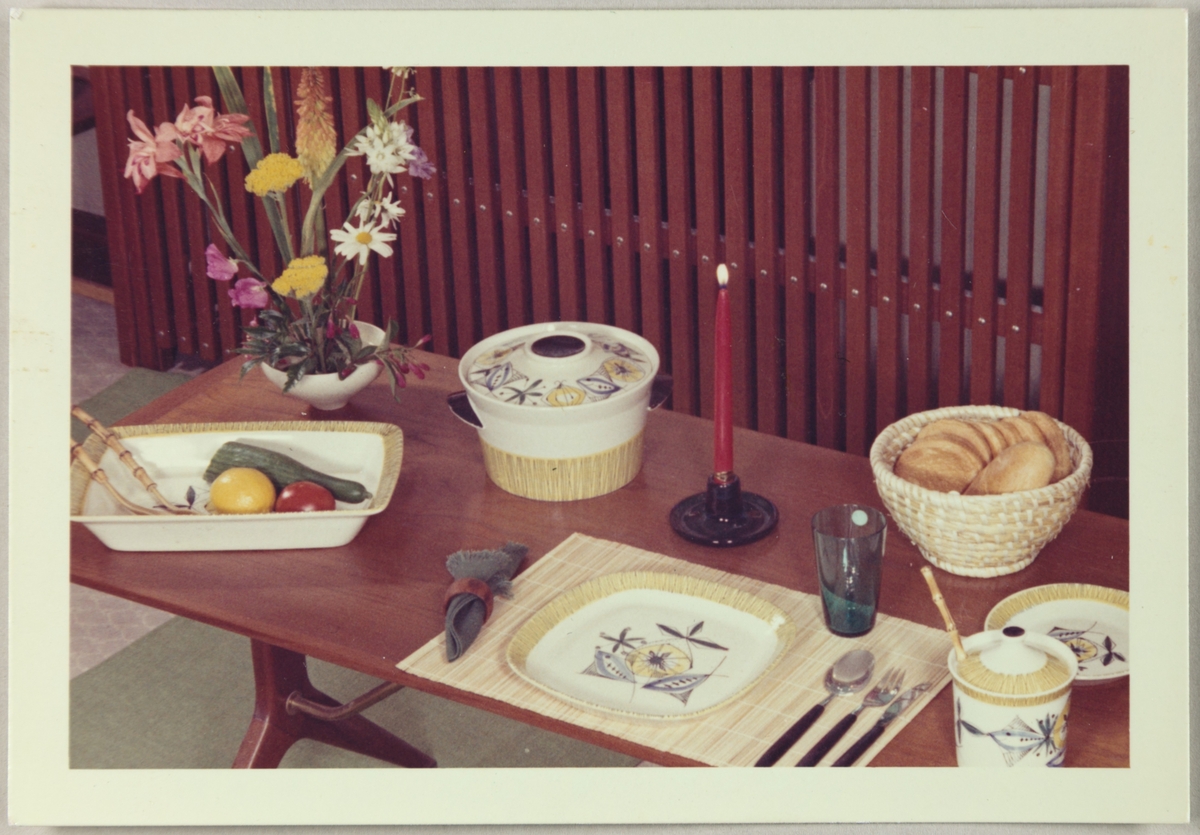 Et bord er dekket med serviset "Bambus" fra Stavangerflint A/S. Avbildede objekter viser en stor og liten tallerken, ildfast gryte og fat, samt en krukke med lokk.
