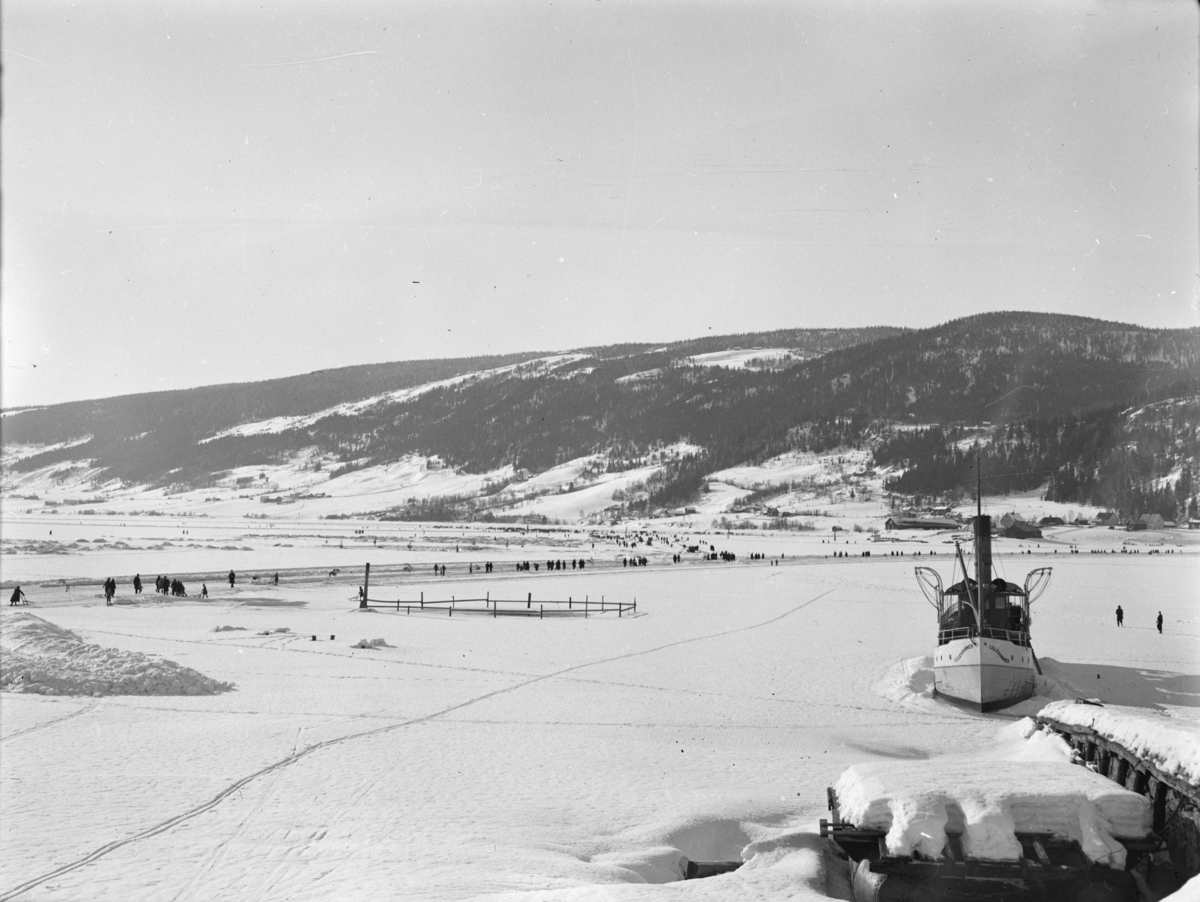 Mjøsa ved Lågendeltaet i Lillehammer dekket av is. Dampskipet Lillehammer ligger ved kai. Mange personer samlet ved brøytet isbane. Vingnes på andre siden.