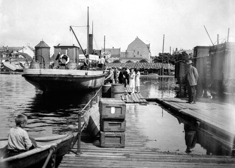 Fotografi av en båt med stavnen mot fotografen, personer som går ombord og en jernbanevogn til høyre i bildet. Strandgata i Hamar skimtes i bakgrunnen.