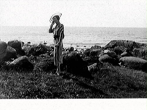En medelålders kvinna klädd i randig klänning och en genomskinlig hatt med två svarta kantcirklar står på stranden vid badplatsen Djupa dräkt i Apelviken. Bild 2 visar närbild på kvinnan och hatten.