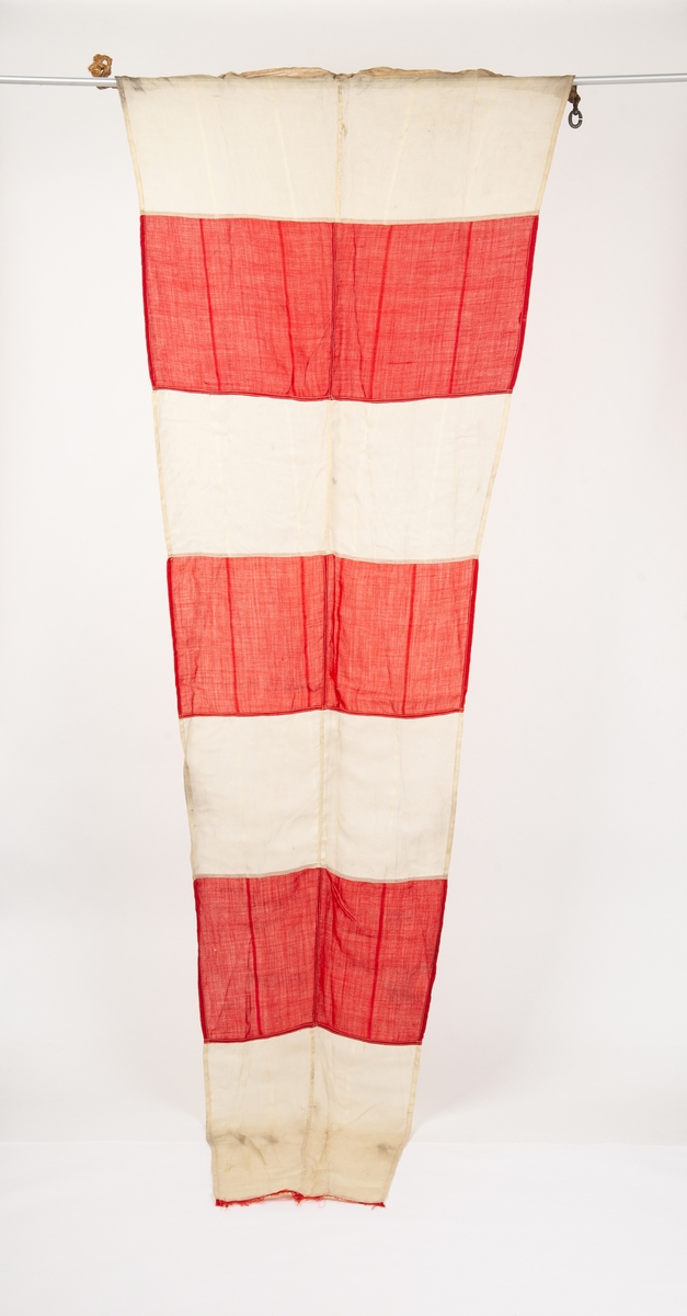 Rektangulært avsmalnende signalflagg. Vekslende røde og hvite vertikale striper. Den ytterste røde stripen har revnet og blitt borte. Forsterkning på innerkanten med tau og opphengskrok.