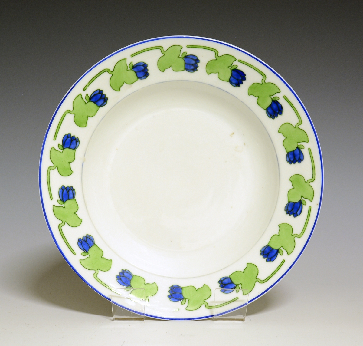 Dyp tallerken i porselen. Hvit glasur. Stilisert bord med blå blomst og grønt blad rundt  fanen.
Modellnr: 15.2

