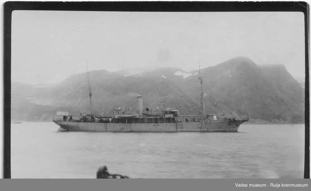 En dampbåt på sjøen i ukjent havn. I bakgrunnen er det høye fjell. Bildet har tilhørt Lars Rygh  fra Hønefoss. Han var med på mineryddingsarbeid i Varangerfjorden 1917 - 1918.