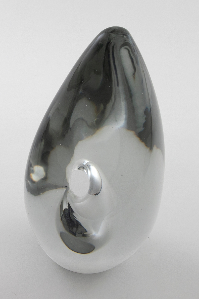 Dråpeformet skulpturell vase med abstrakt utforming i massivt klart glass. Et elipseformet hulerom i nedre del med liten sirkulær åpning. Bunnen er planslipt.