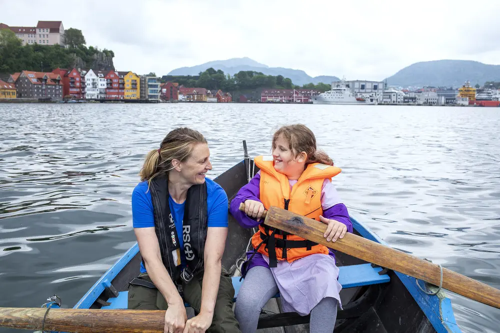 En voksen dame og en jente, begge med redningsvester, ror hver sin åre i en robåt på vannet ved Norges Fiskerimuseum