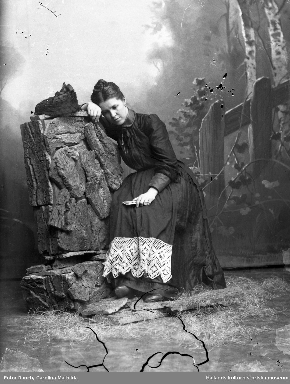 Arrangerat fotografi (utan år) av Mathilda med ett fotografi i handen. Illustration av en kvinna som sitter i ett skogsbryn och är sorgsen över den älskade på bilden. Hon bär en sannolikt svart klänning med bred spets nedtill.