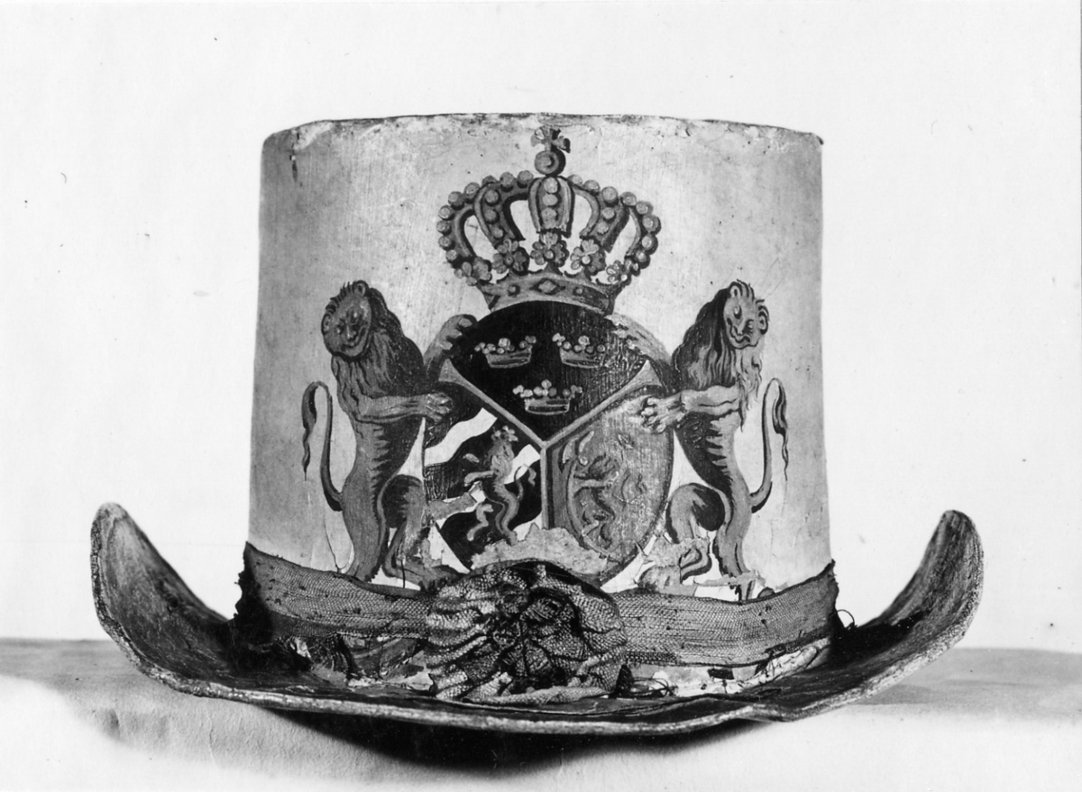 Hatt tillverkad av papp som målats med en crémegul färg. Framtill försedd med svensk-norska riksvapnet i färg. Foder av bomullstyg, 115 mm brett och dragsko.