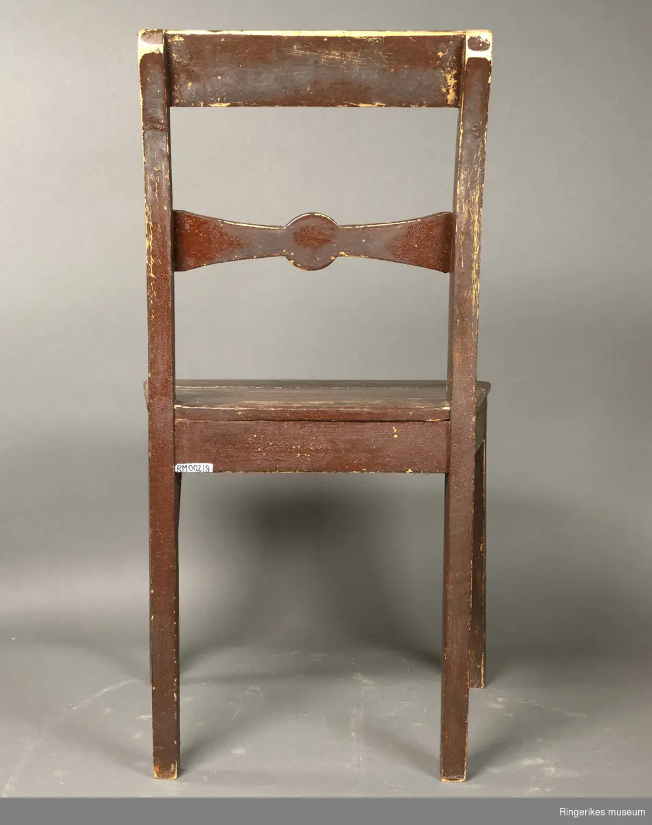 Brunmalt empire stol i furu. Stolen har ikke sprosser mellom stolbenene. Enkelt toppstykke og ryggbrett med sløyfeform. Sargen er bredere i front enn bak og har en svak bue i front.