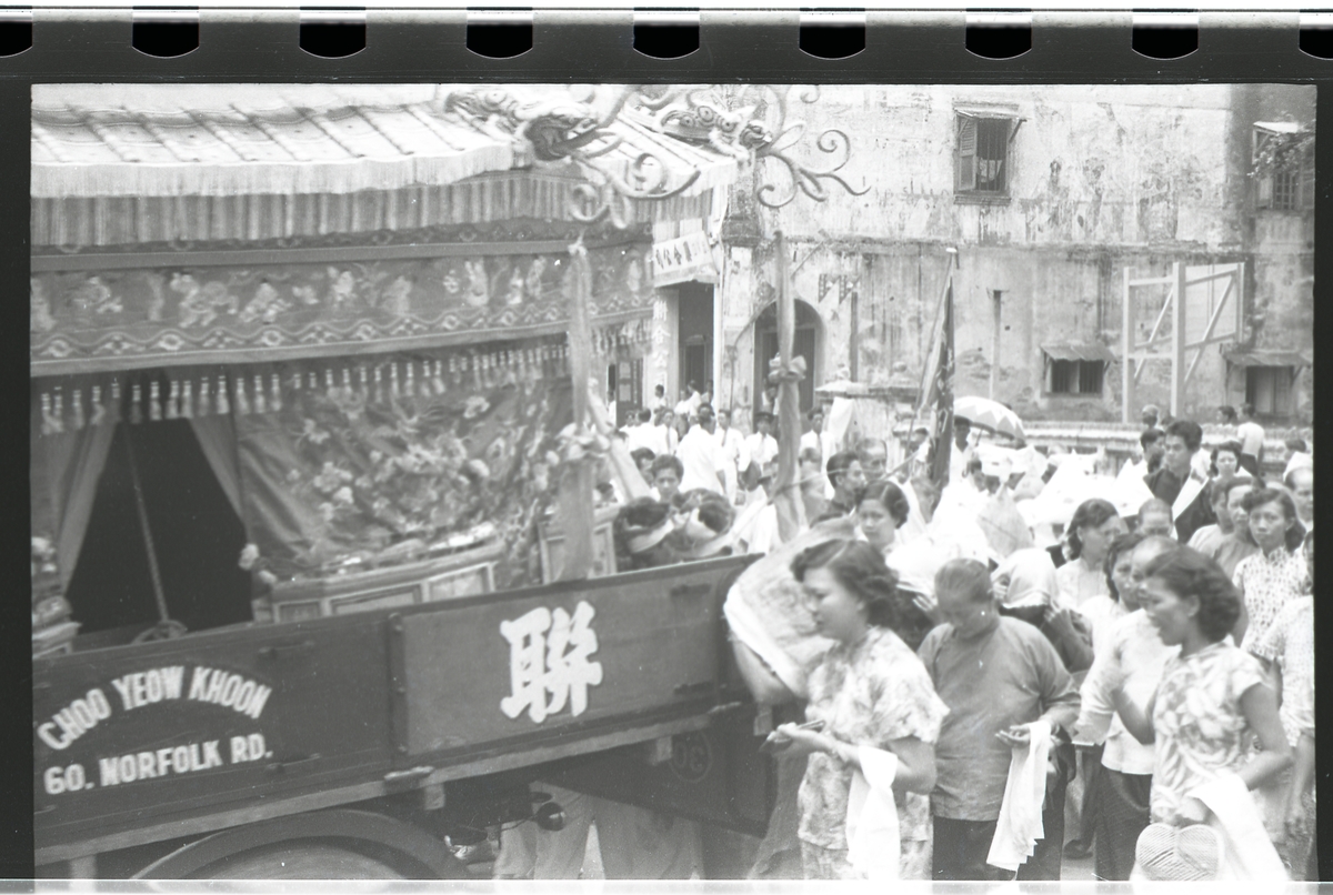 Seremoniell begravelsesprosesjon gjennom gate i Singapore. Bil eller vogn merket med "Choo Yeow Khoon 60. Norfolk Rd." og det kinesiske tegn for "Forent".