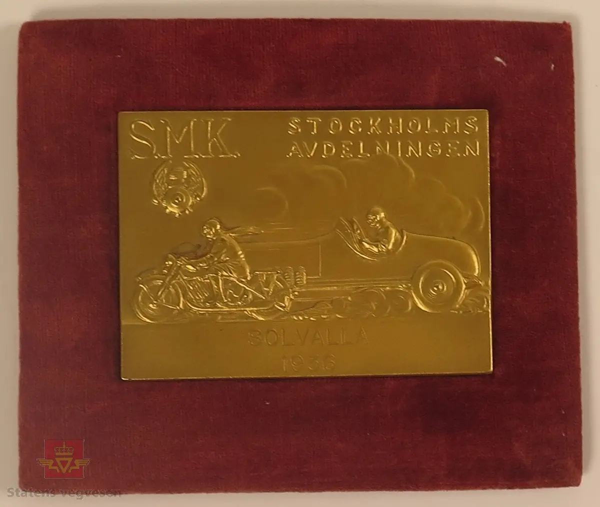 Plakett laget av metall i gull farge, rammet av rødt velour stoff. På plaketten er det utformet en bil og motorsykkel som tilsynelatende kjører om kapp.