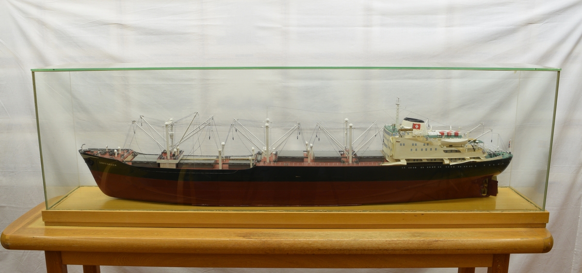 Båtmodell med glasslokk i som kan løftes av plate med påmontert modell. Plate og listverk i lakkert lyst tre. Modell i tre, plast og metall.