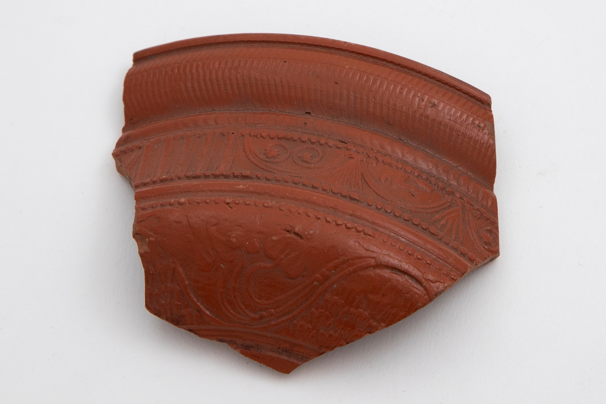 Fragment av arrentinsk vase i terrakotta, dekorert med rødfarget begitning. På yttersiden dekor i lavt relieff som skal forestille rankemotiv, samt rektangulære felt med drapperier. Øvre del har en rillet kant.