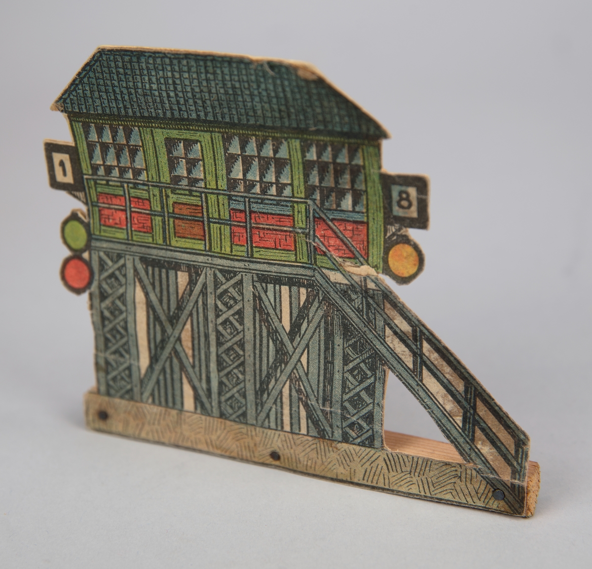 Pappersmodell föreställande ställverkbyggnad i grönt, grått och rött. Pappersmodellen fäst i smal träpinne med tre spikar.