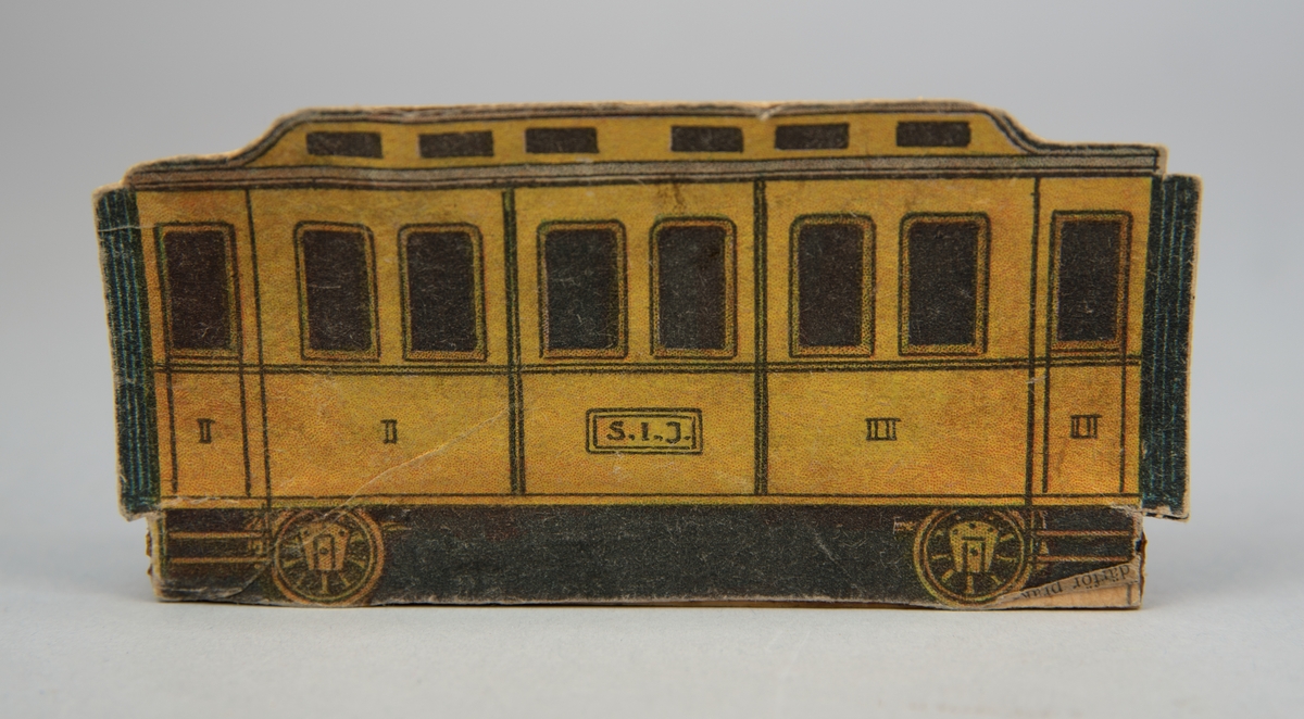 Pappersmodell föreställande gul personvagn. På vagnen finns skylt märkt "S.I.J.". Pappersmodellen fastlimmad på smal träpinne.