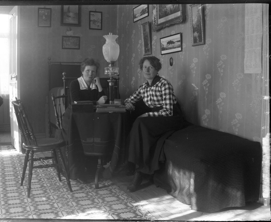 Två kvinnor sitter vid ett bord med en hög fotogenlampa.