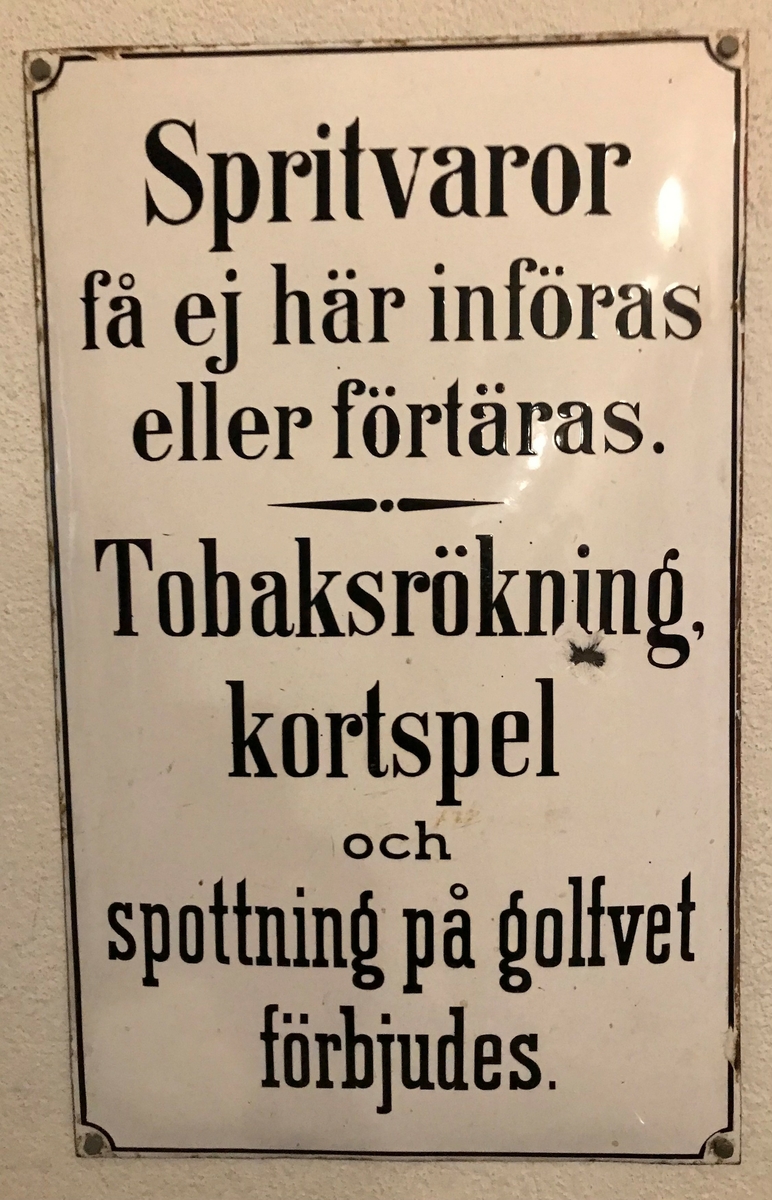 Skylt, rektangulär, vitemaljerad, med text i svart: "Spritvaror få ej här införas eller förtäras. Tobaksrökning kortspel och spottning på golvet förbjudes."