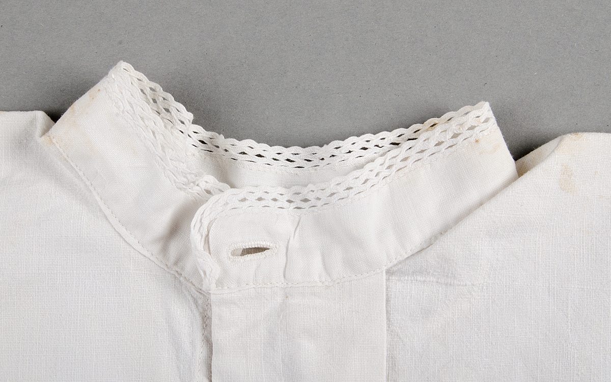 Skjorta av vit bomullslärft. Rund hals och ärmar genombrutet band av bomull. Knäpps fram med en knapp i ståndkragen. Fällsömmar. Maskinsydd.
