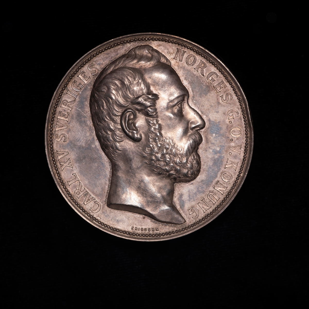 Sølvmedalje fra utstilling i Stockholm 1866. På adversen portrett mot høyre av kong Carl 15. På reversen kvinnelig guddom deler ut laurbærkranser til fire sittende kvinner.
