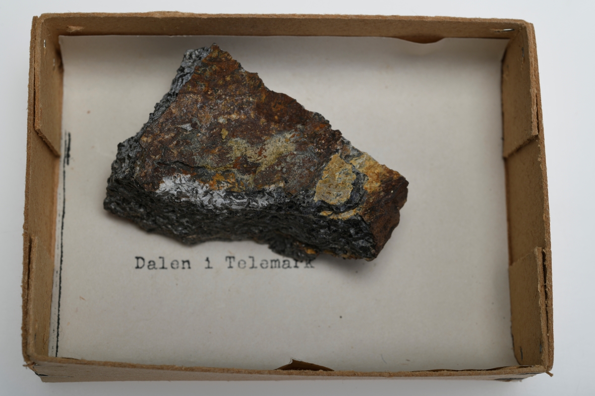 Et stykke kaliglimmer (også kjent som kråkesølv) fra Dalen i Telemark. Over- og underside er brun med brungule flekker. Sidene er svarte og har en sølvglinsende overflate. 
Del av steinsamlingen som inngår i skolesamlingen.