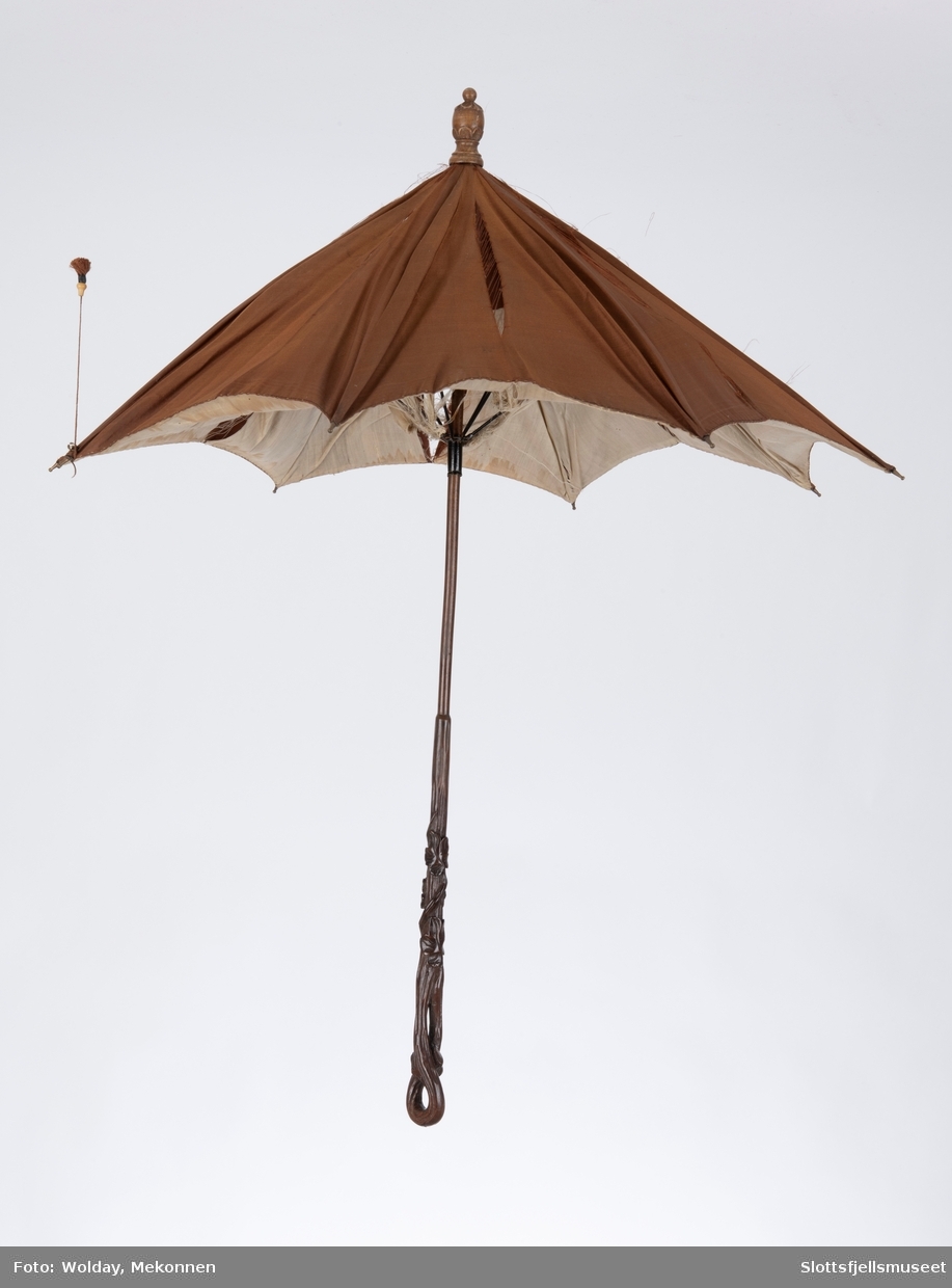 Parasoll med  8 spiler av metall. Parasoll har en utskåret treskaft og er trukket utvendig med brunt silke, innvendig med lys silke. Ved en av spilene er en snor med dusk montert for å holde spilene sammen når parasoll er lukket. 