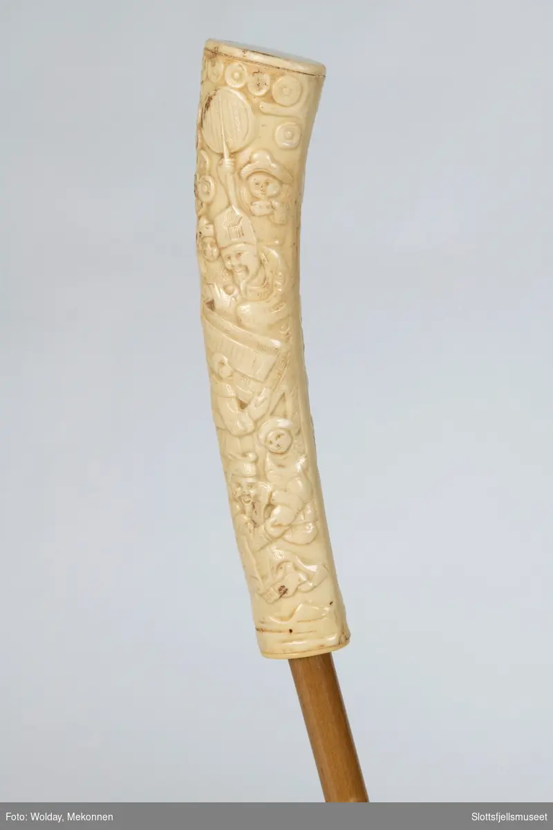 Parasoll med 12 spiler, trukket med lyst bomullsstoff. Treskaft som ender i en håndtak av bein. Svakt buet håndtak med utskjæringer i asiatisk stil. 