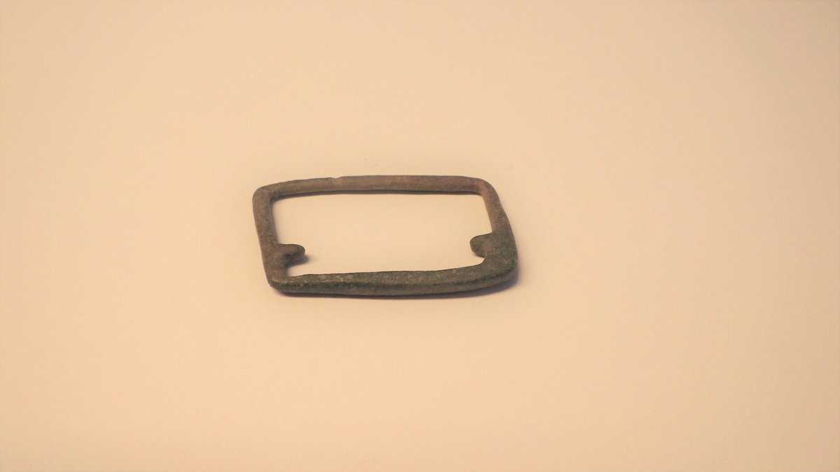 Rektangulær og rombeformet enkel metallspenne til bruk på et belte. Spennen er av en type som ikke har hatt torn,men har to tagger inni rammen. Beltet ble festet ved å trekkes gjennom spennen.