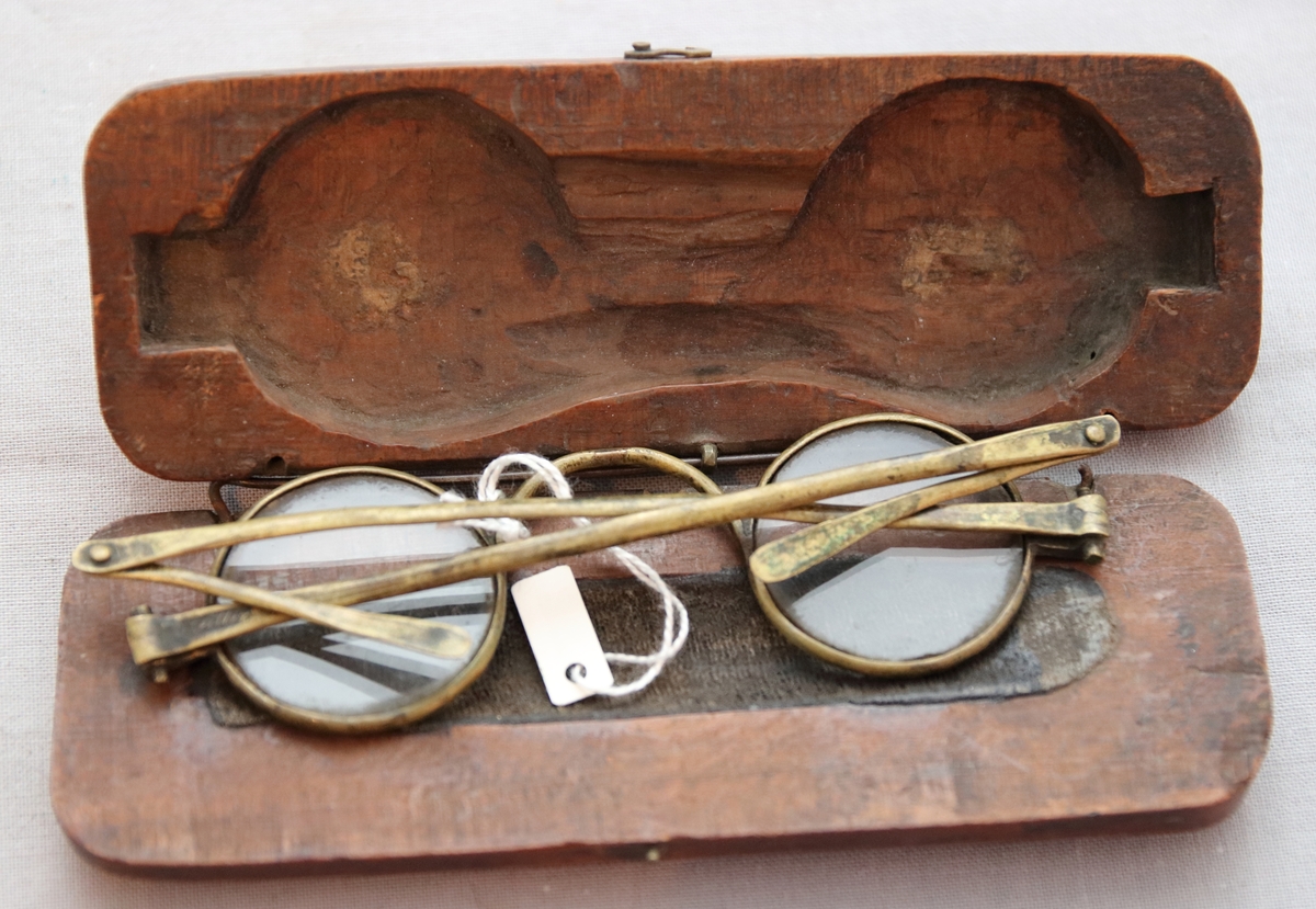 Glasögon i fodral. Fodralet består av två halvor i solida träbitar som sedan är urgröpta så att glasögonen får plats och har gångjärn i metall. Glasögonen har bågar av mässing och runda glas med utfällbara skalmar.