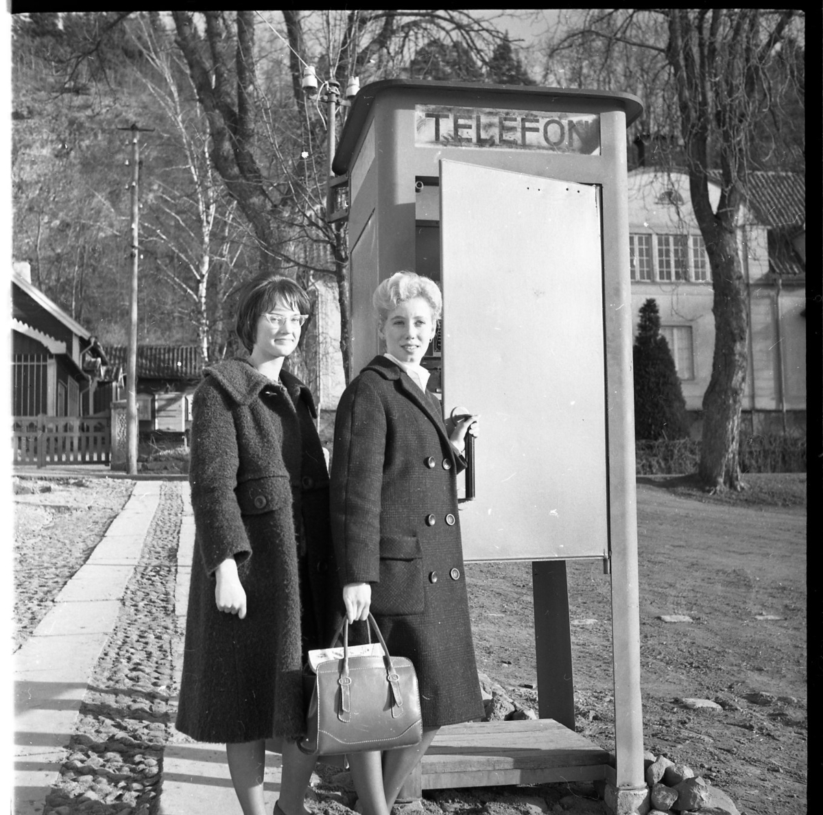Två kvinnor står utanför en telefonkiosk. De bär kappor och har tuperat hår. En av dem bär en handväska och den andra bär ett par glasögon som är typiska för 1960-talet. I bakgrunden syns riksinternatskolan Grennaskolan.