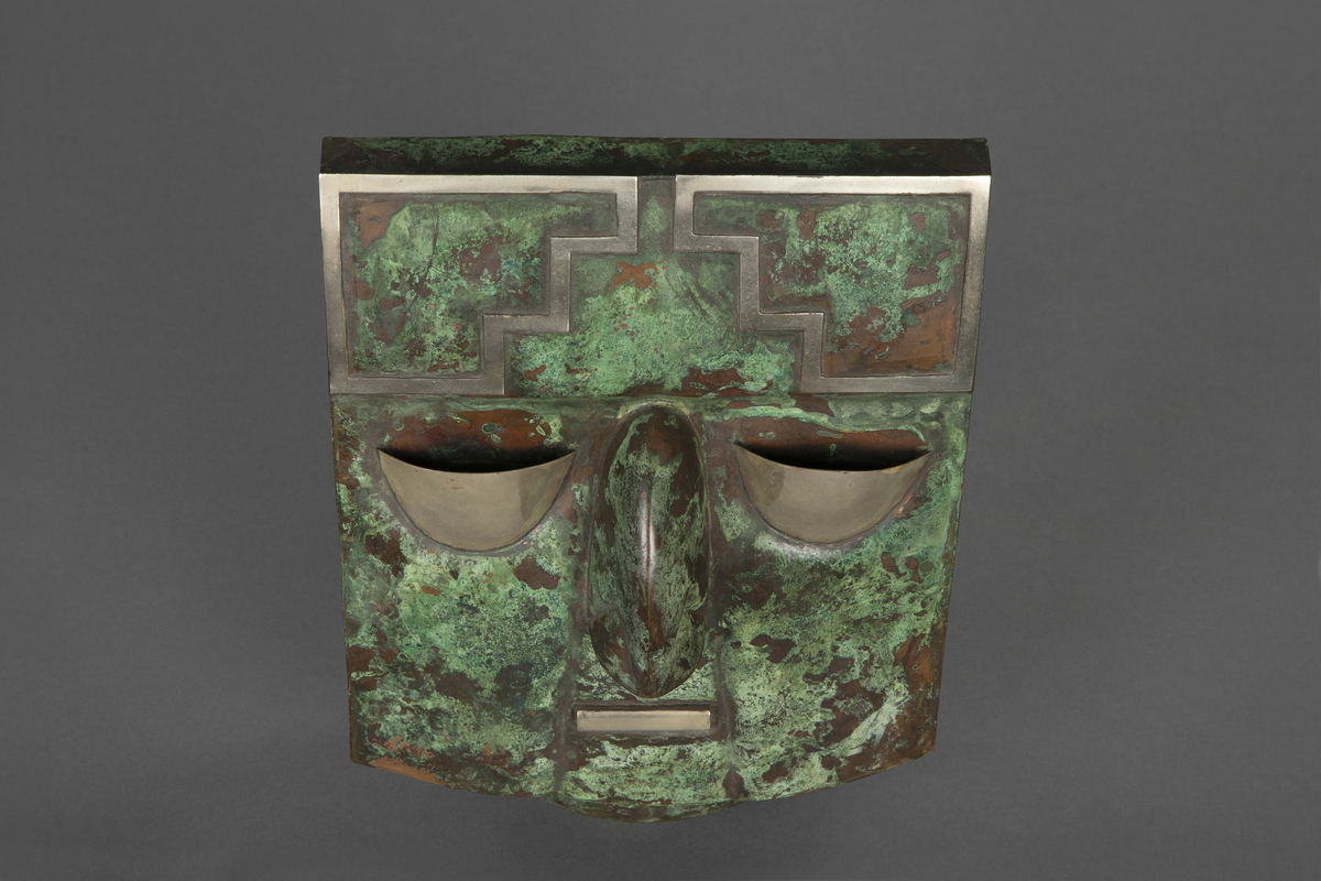 Maske av metallplater i kobber med messingdetaljer. Er med oppheng montert på baksiden. 