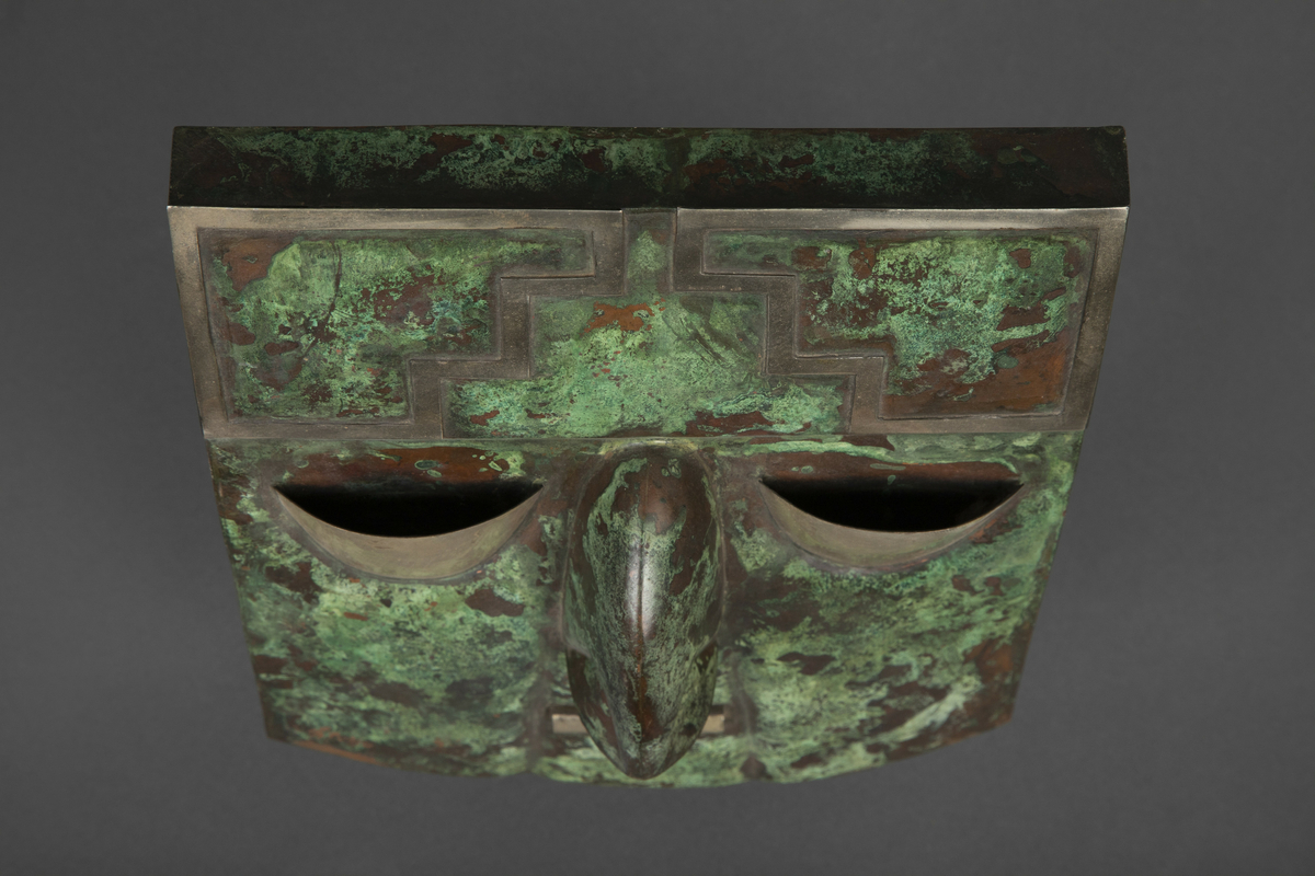 Maske av metallplater i kobber med messingdetaljer. Er med oppheng montert på baksiden. 