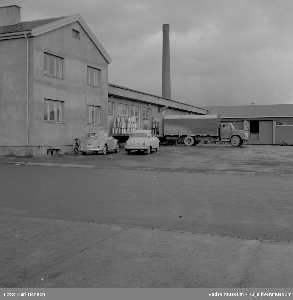A/L Aust-Finnmark Meieri, 1962 i Vadsø. Bildet er tatt mot nordøst. En lastebil er rygget inn til bygningen for av/pålasting og to personbiler står parkert på sydsiden av bygningen.
Arbeidet med å få meieri til Vadsø ble påbegynt før krigen.  Ønsket var å få et meieri som kunne dekke Nord-Varanger-Tanadistriktet.  Andelslaget A/L Aust-Finnmark Meieri ble stiftet 28.juni 1947 i Nyborg.  
Aust- Finnmark Meieri ble oppført i Tollbugata i 1949; arkitekt Monrad Heggen m hovedentreprenør Brdr. Aronsen.
Oppstart skulle finne sted ca 1. oktober 1949, og i mai ble det ansatt en meieriarbeider; Richard Korbi fra Vadsø.
Kilde: "A.L Aust-Finnmark Meieri 25 års jubileum 1949-1974; styreformann Ivar Johnsen, Tana mai 1974"