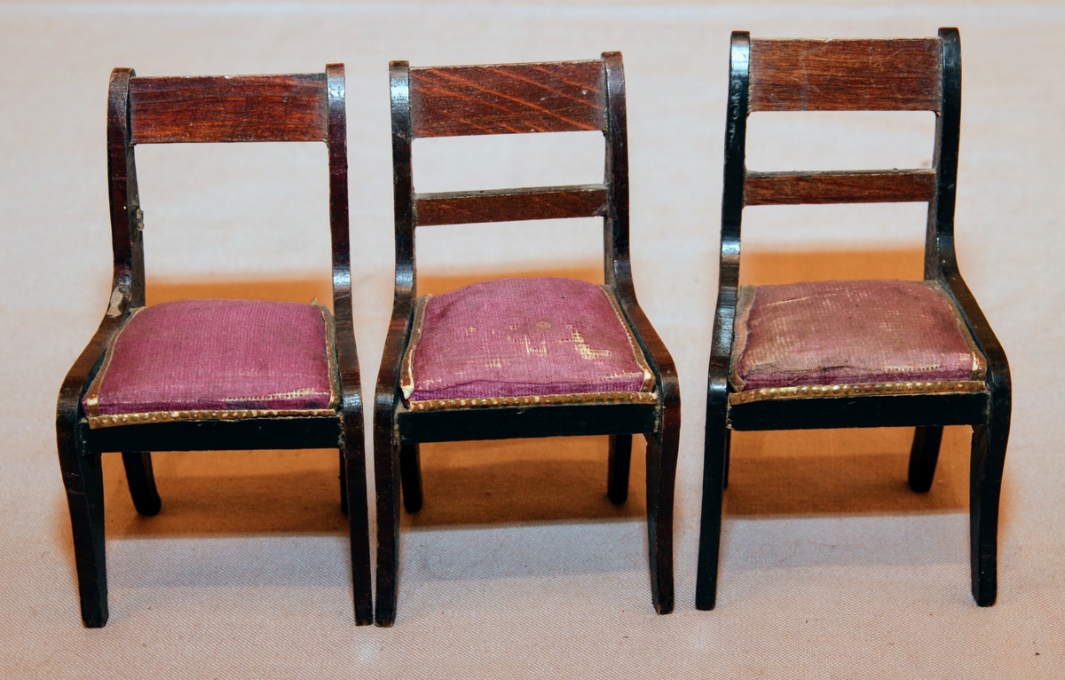 Tre stolar till dockskåp från mitten av 1800-talet i empirestil. I mörkbrunt trä med lila stoppade sitsar och guldiga pärlbårdar kring sitsarna. Med två tvärslåar i ryggen (en saknas på en av stolarna).