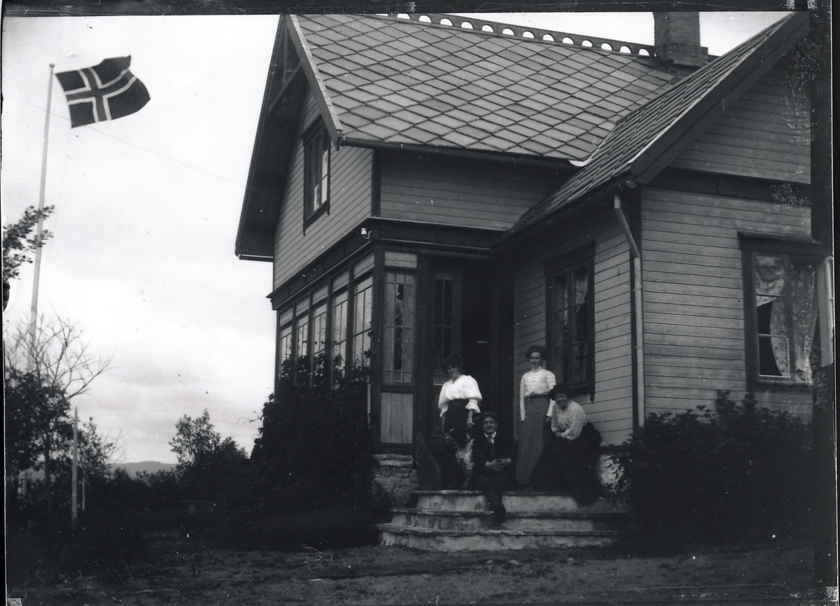 Tre kvinner og en mann på verandatrapp utenfor stort bolighus med preg av dragestil. Ene kvinnen holder en hund. Flaggstang med norsk flagg til venstre.