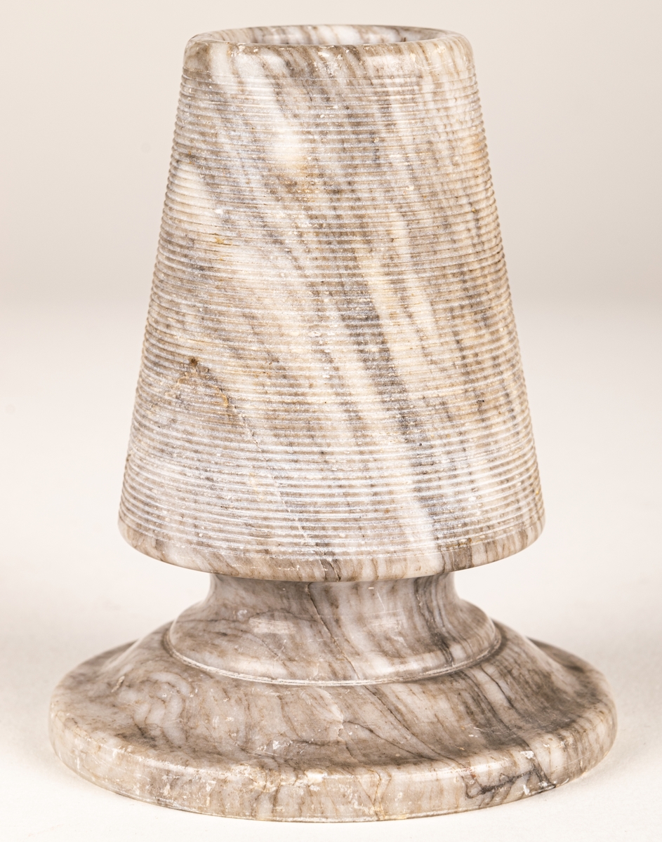 Tändsticksställ, av gråvit marmor, i form av stympad kon på rund fot. Hållaren ovanpå räfflad.