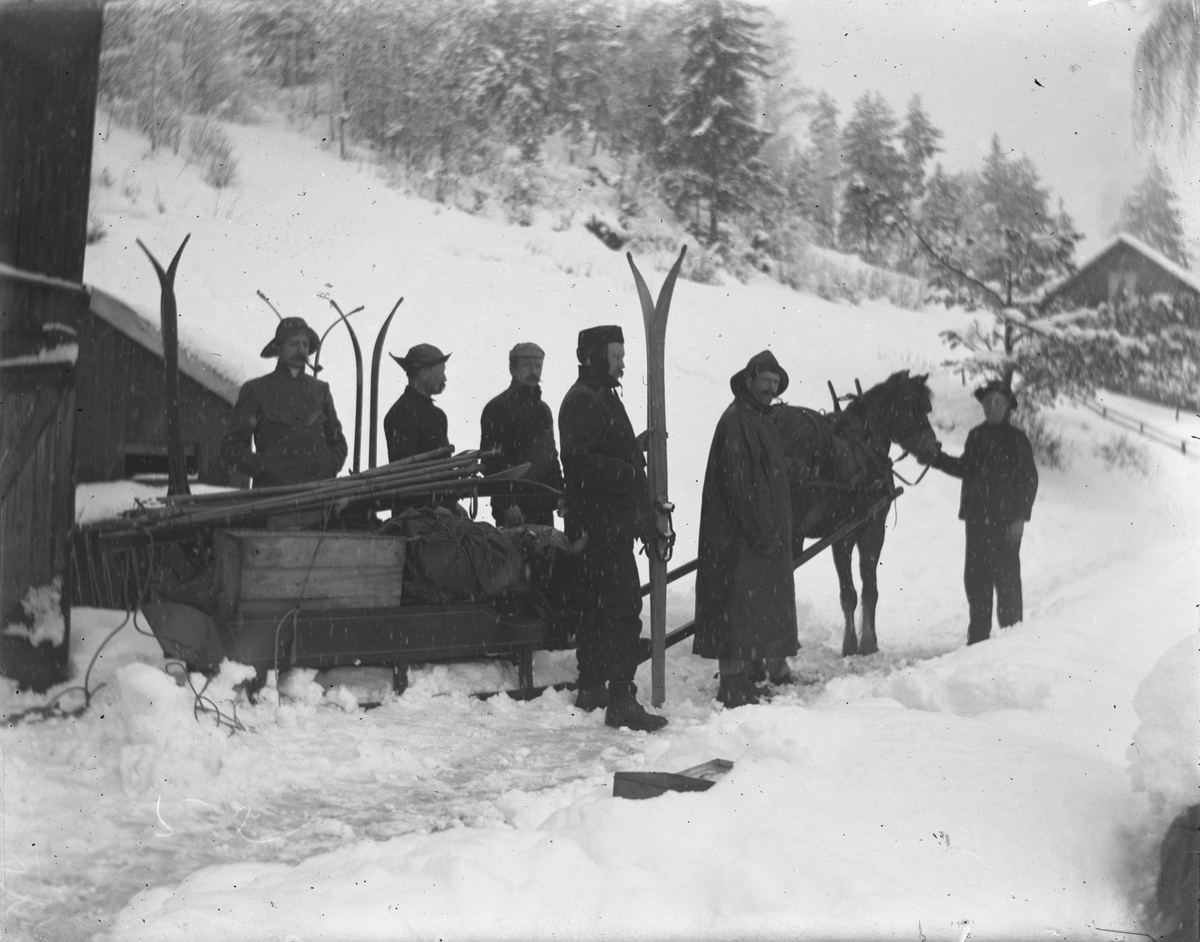 Hest og slede, seks menn, noen med ski på en gård om vinteren, snøvær?