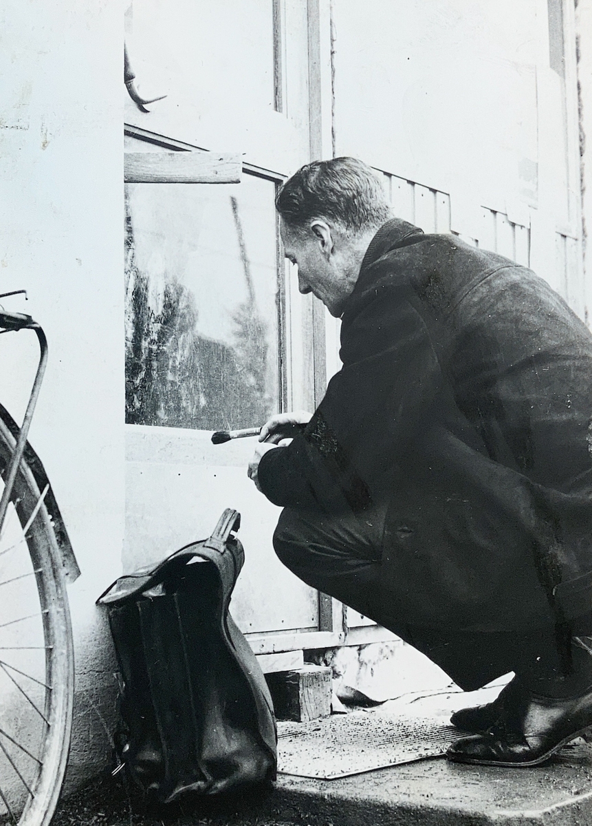 En mann sitter på huk foran en glassrute med en pensel i hånden. Veske, sykkel.