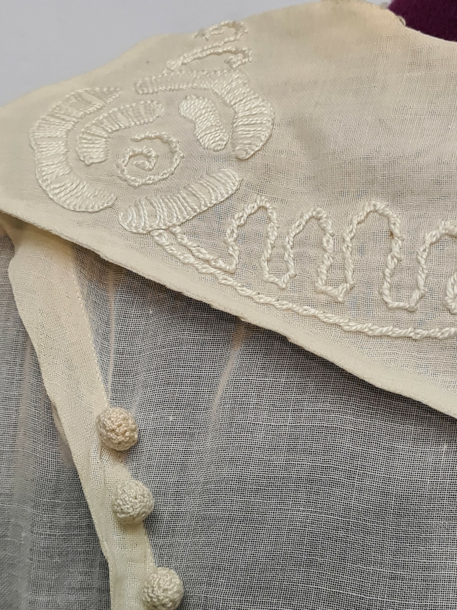 V-ringad klänning med brodyr i vitt.  Virkade runda knappar, liten broderad krage bak. Sydd till bröllop 1911. Från Sörmland.