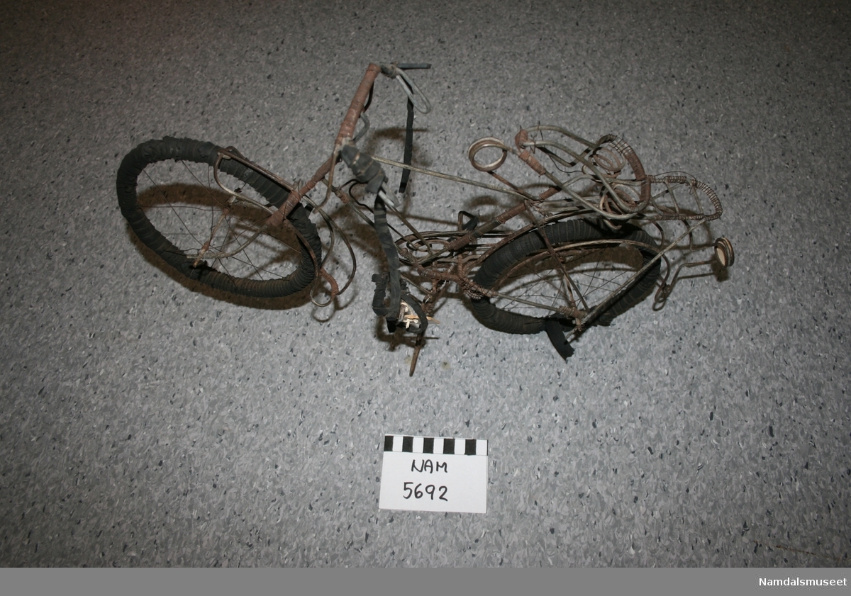 Teknikk: Ståltråd bøyd til som rammen til en vanlig sykkel med sete, ratt osv. Hjulene surret med gummi. Ratt og pedaler surret med gummi.
