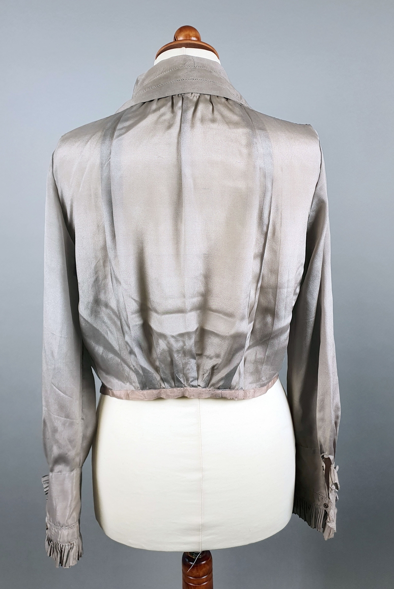 Gråbrun silkebluse med jakkeslag med rysjer langs kanten og over brystet. Blusen lukkes med trykknapper. Lange ermer med mansjetter med knapper og rysjekant.