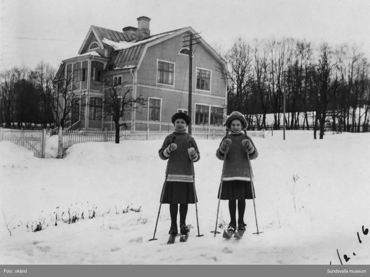 Två skidåkande flickor framför ett vackert bostadshus med glasverandor. Ur ett album från Harv, Attmar.