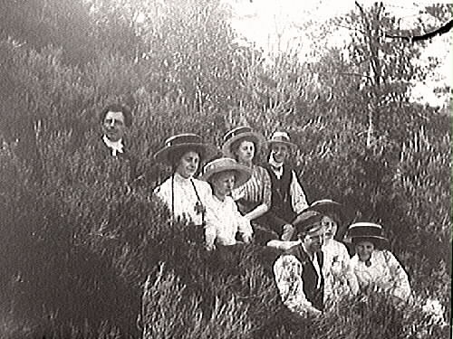 Utflykt. Ett sällskap män och hattbeklädda kvinnor sitter i en skogsbacke.