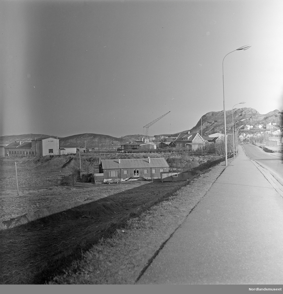 Bodø, bygninger, boligen til Bodø Teglverk. En bil står parkert til venstre foran boligen. Brakke til høyre foran boligen. Bebyggelse lenger bak til høyre. En kran ses midt på bildet i bakgrunnen. Bilde 2 er en positivkopi av et utsnitt av bildet.
