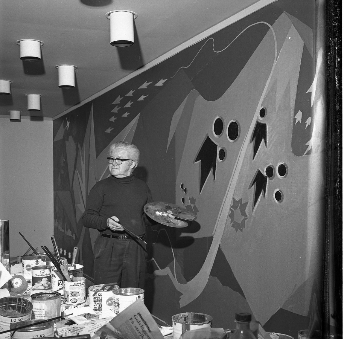 Georg Trapp arbetar vid en stor väggmålning. Färgburkar står på en bänk framför honom.