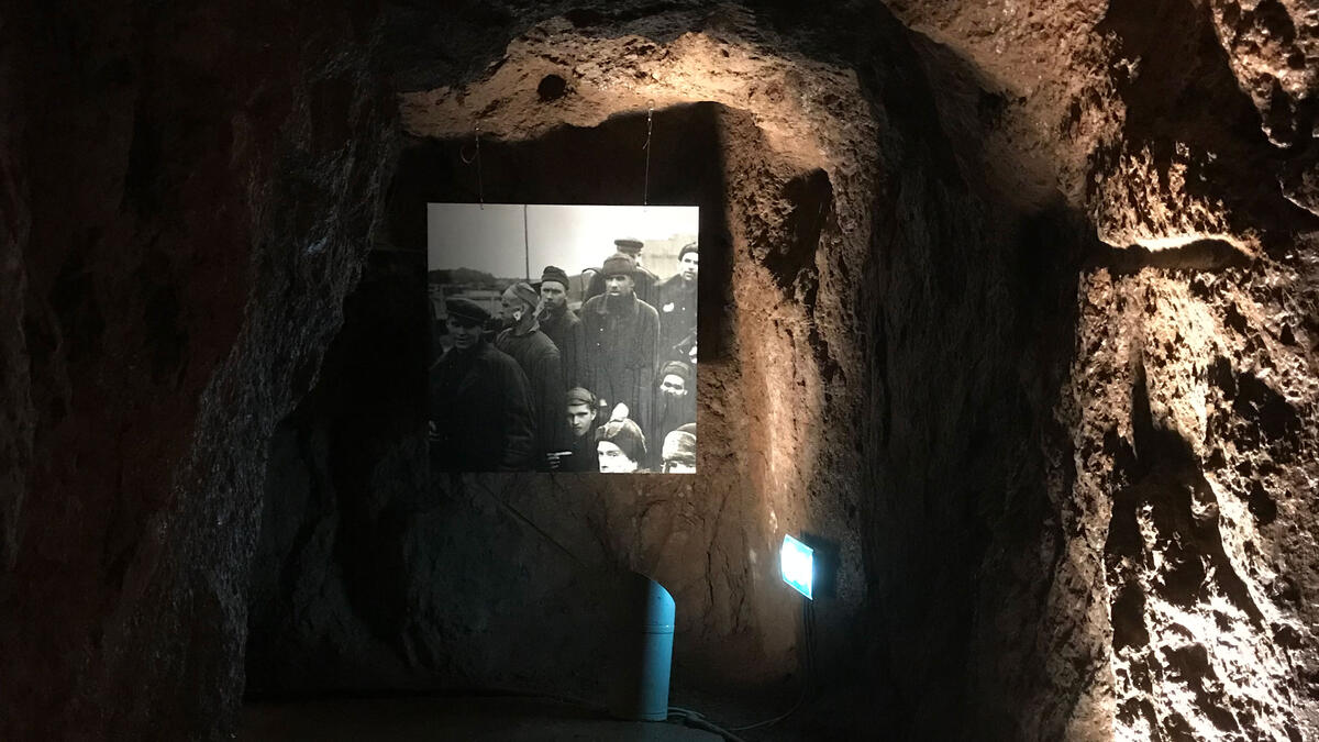Vi ser et sort-hvit-fotografi som henger på en ruglete steinvegg i et mørkt rom, det vil si inne i en hule. (Foto/Photo)