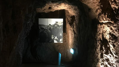 Vi ser et sort-hvit-fotografi som henger på en ruglete steinvegg i et mørkt rom, det vil si inne i en hule.. Foto/Photo