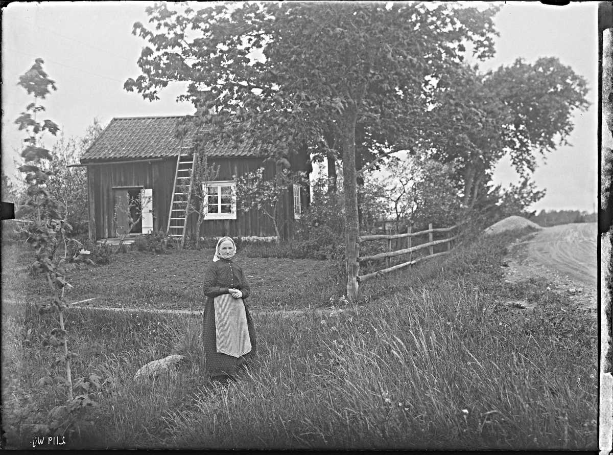 Fotografering beställd av Johansson. Föreställer sannolikt änkan och lägenhetsägaren Maria Josefina Johansson, född Svärd 1866-02-28 i Tillinge, död 1955-10-31 i Tillinge. I bakgrunden syns då gamla soldatstugan där hon bodde.
