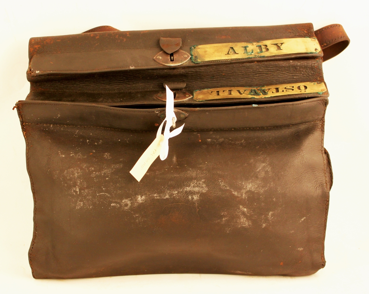 Lösväska i läder med bärrem och låsinrättning. Två fack med inskrift utskurna i mässingsplåtar: Alby respektive Östavall. Varje fack är låsbart separat.